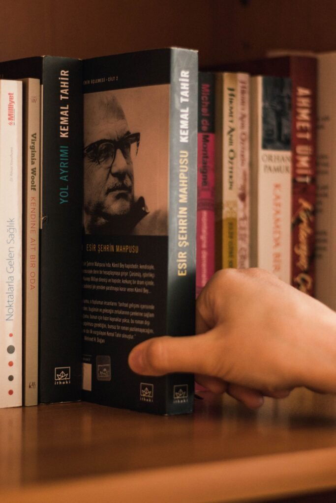 Crop anonymous person selecting modern book with photo of author in eyeglasses with text on back cover near assorted literature on wooden bookshelf in house